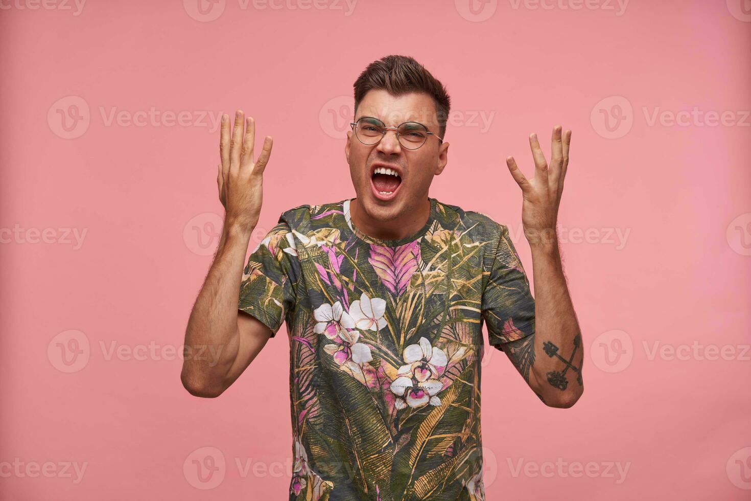 verständnislos verwirrt jung Mann mit Hände oben Stehen Über Rosa Hintergrund, tragen T-Shirt mit Blumen- drucken und Gläser, suchen verwirrt foto