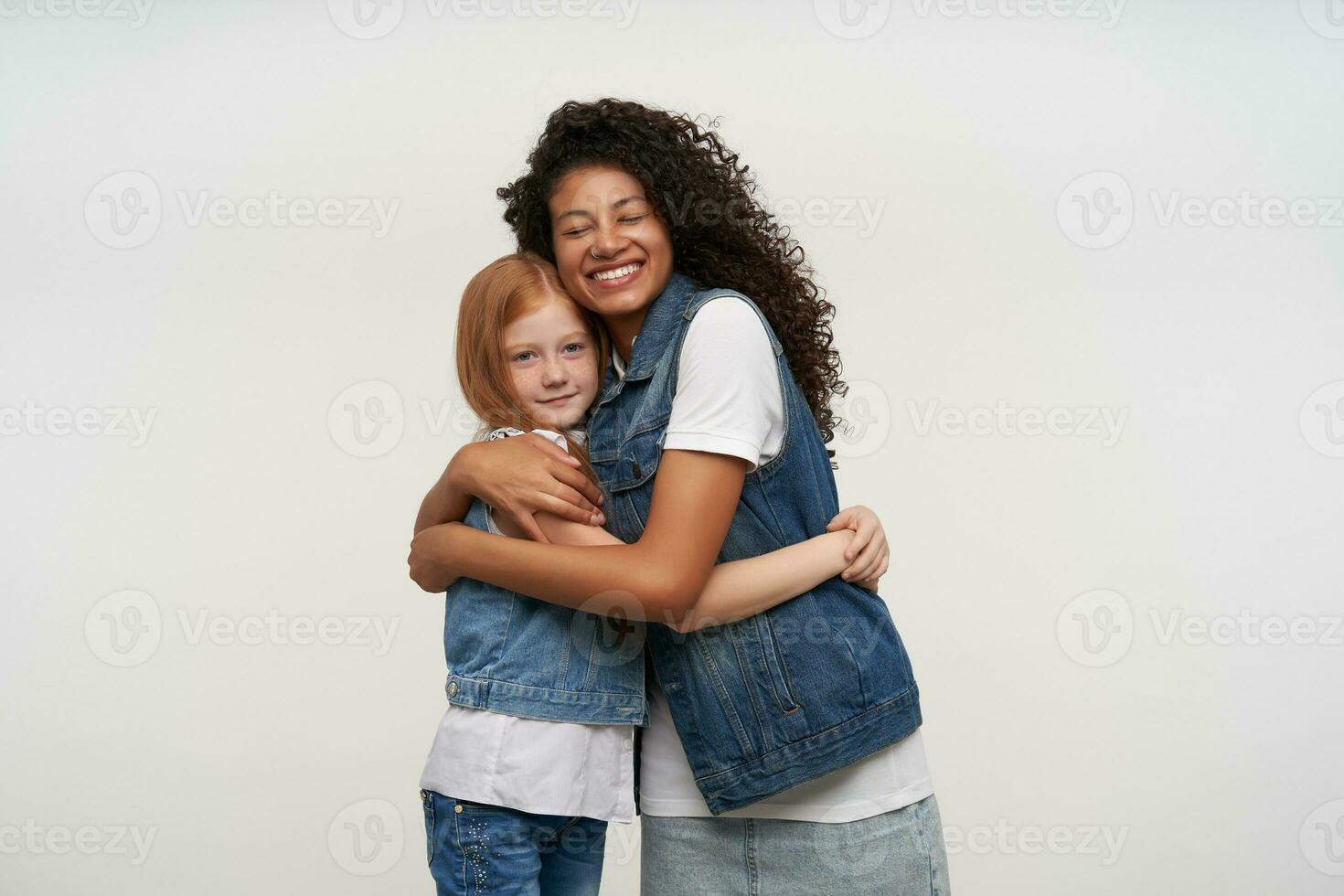 Studio Schuss von glücklich dunkel gehäutet lockig Brünette Frau umarmen vorsichtig ziemlich lang behaart foxy Mädchen und behalten ihr Augen geschlossen, isoliert Über Weiß Hintergrund foto