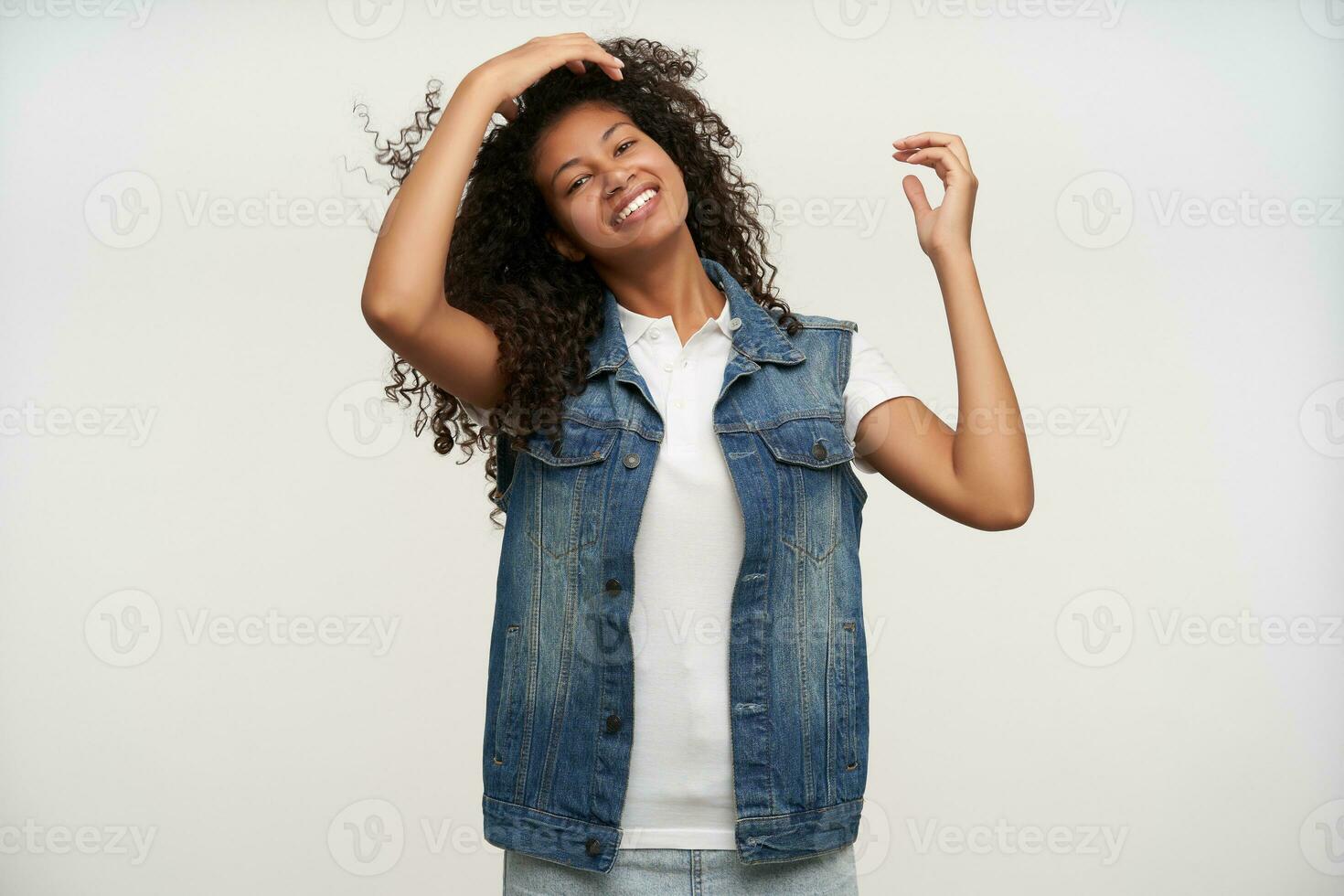 Studio Schuss von ziemlich Brünette lockig Frau mit dunkel Haut begradigen ihr Haar mit angehoben Hand und suchen fröhlich zu Kamera, gekleidet im Jeans Weste und Weiß Hemd Über Weiß Hintergrund foto