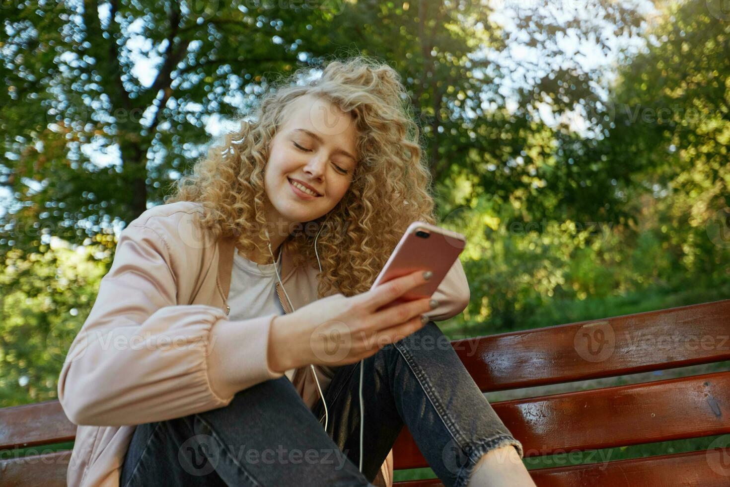 jung schön Mädchen blond mit lockig Haar sitzt mit ihr Beine auf ein Park Bank, hört zu zu ihr Liebling Musik- im Kopfhörer mit Augen geschlossen von Vergnügen, tragen ein Rosa Jacke und schwarz Pfannen foto