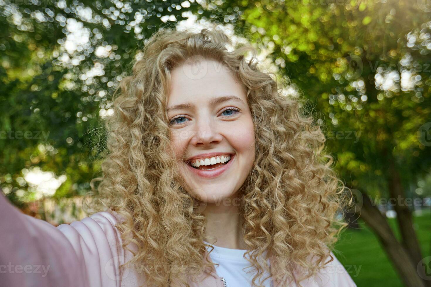 attraktiv Mädchen mit lockig Haar und Blau charmant Augen, angenehm lächelnd, suchen beim Kamera, Herstellung Selfie, Natur Hintergrund foto