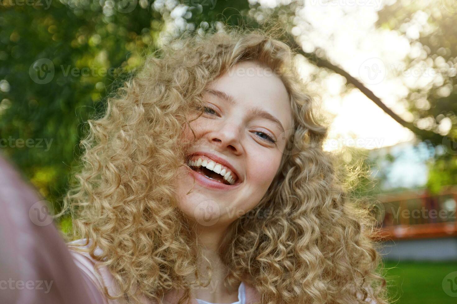 jung schön komisch cool Mädchen blond mit lockig Haar, weit lächelnd und suchen in das Kamera, Herstellung Selfie, Kopf gekippt zu das Seite, Natur Hintergrund foto