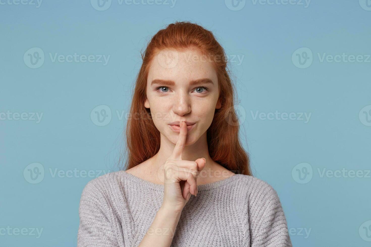 Porträt von nett heiter rothaarig jung weiblich zeigt ein Geste von Stille, hält Finger beim ihr Mund, scherzhaft sieht aus beim das Kamera, fragt zu behalten ein Geheimnis, Geheimnis, vorbei Blau Hintergrund. foto