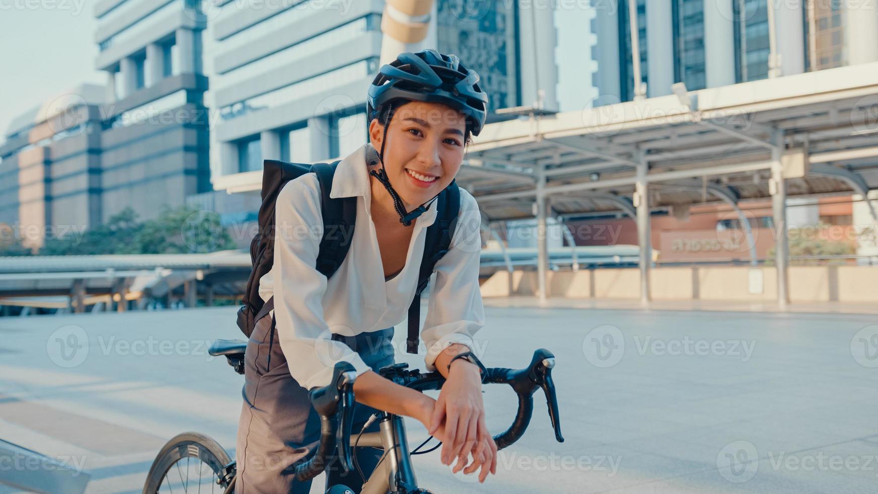 Asiatische Geschäftsfrau geht am Bürostand zur Arbeit und trägt lächelnd Rucksack Blick in die Kamera mit Fahrrad auf der Straße rund um das Gebäude in einer Stadt. Fahrradpendeln, Pendeln mit dem Fahrrad, Geschäftspendlerkonzept. foto