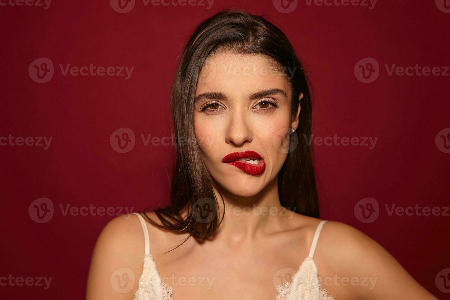 Studio Foto von jung ziemlich Brünette Frau mit Abend bilden beißen ihr Unterlippe und Schielen ihr Augen während suchen beim Kamera, isoliert Über Burgund Hintergrund