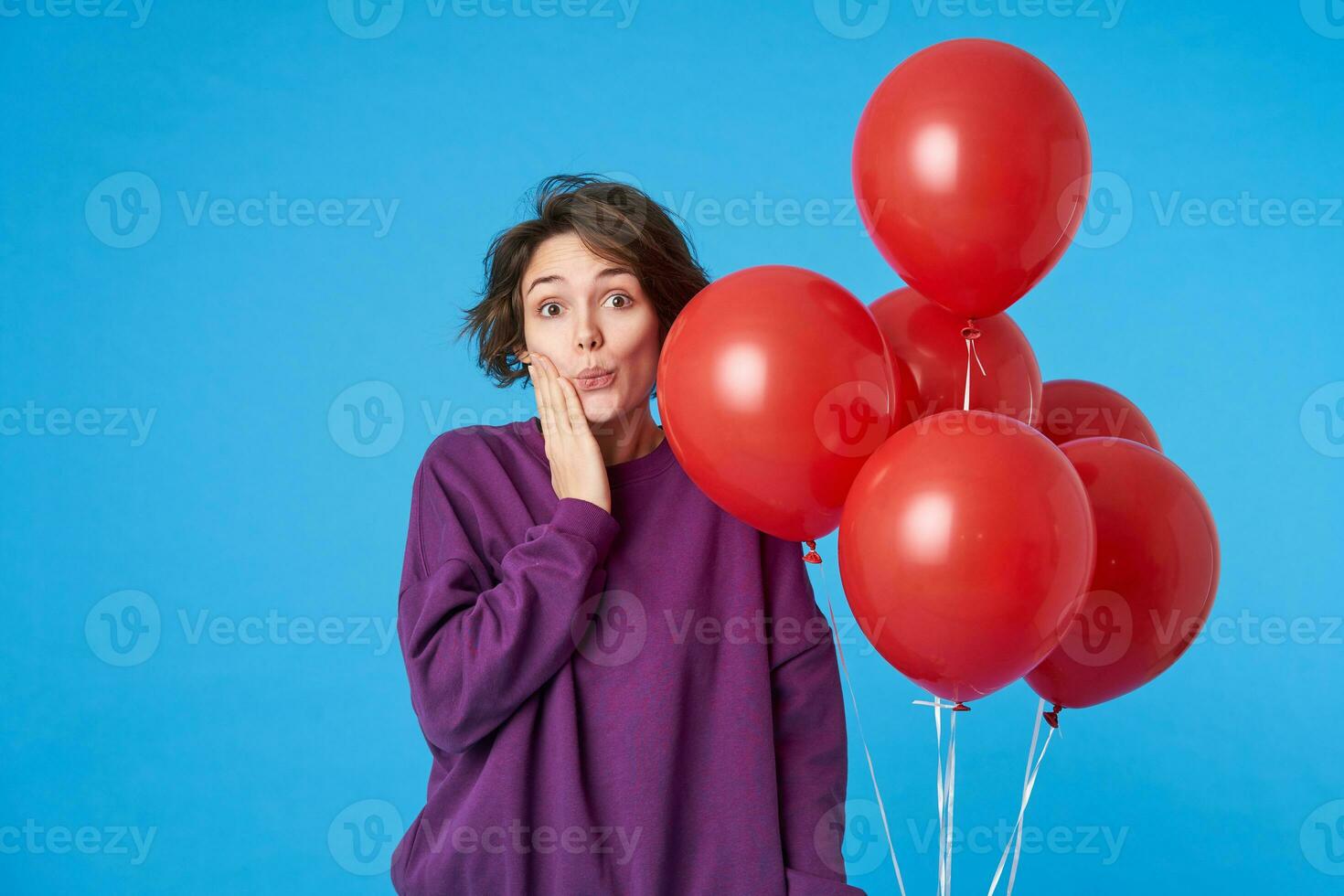 verwirrt jung ziemlich lockig Brünette Frau mit beiläufig Frisur berühren ihr Gesicht mit angehoben Hand und suchen wundernd beim Kamera, Stehen Über Blau Hintergrund foto