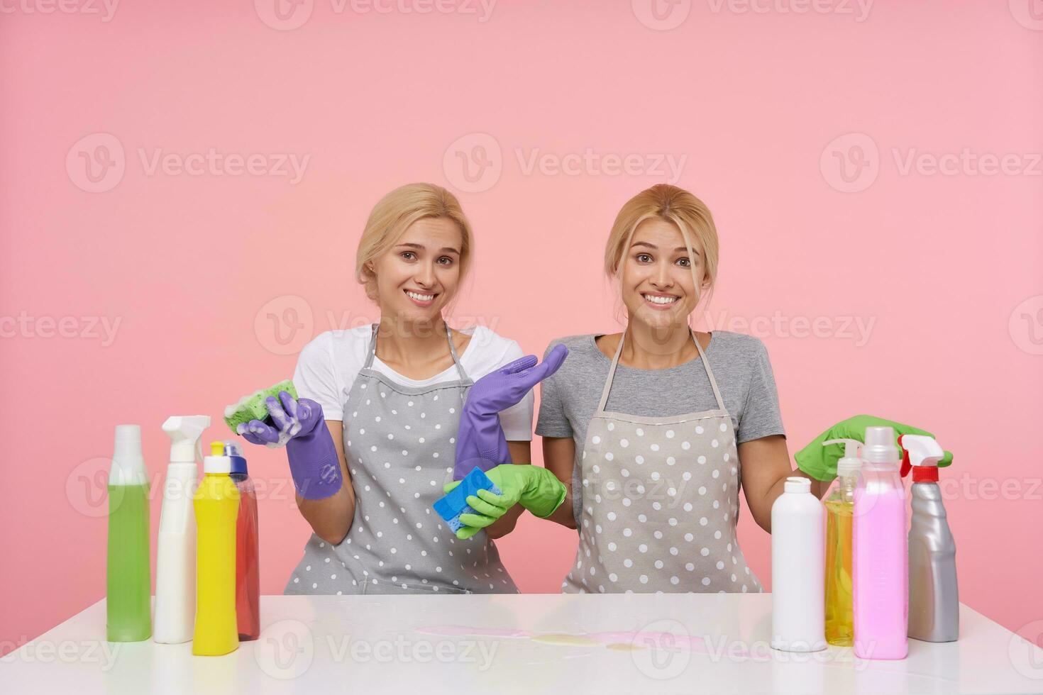 heiter jung blond Damen gekleidet im Arbeiten Kleider und Gummi Handschuhe umarmt mit angehoben Hände und lächelnd gern beim Kamera, Sitzung Über Rosa Hintergrund foto