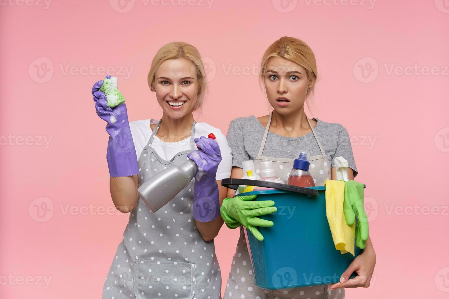 Studio Foto von jung braunäugig blond Frauen gekleidet im beiläufig T-Shirts und Polka Punkt Schürzen Herstellung Frühling Reinigung zusammen und halten Waschmittel, isoliert Über Rosa Hintergrund