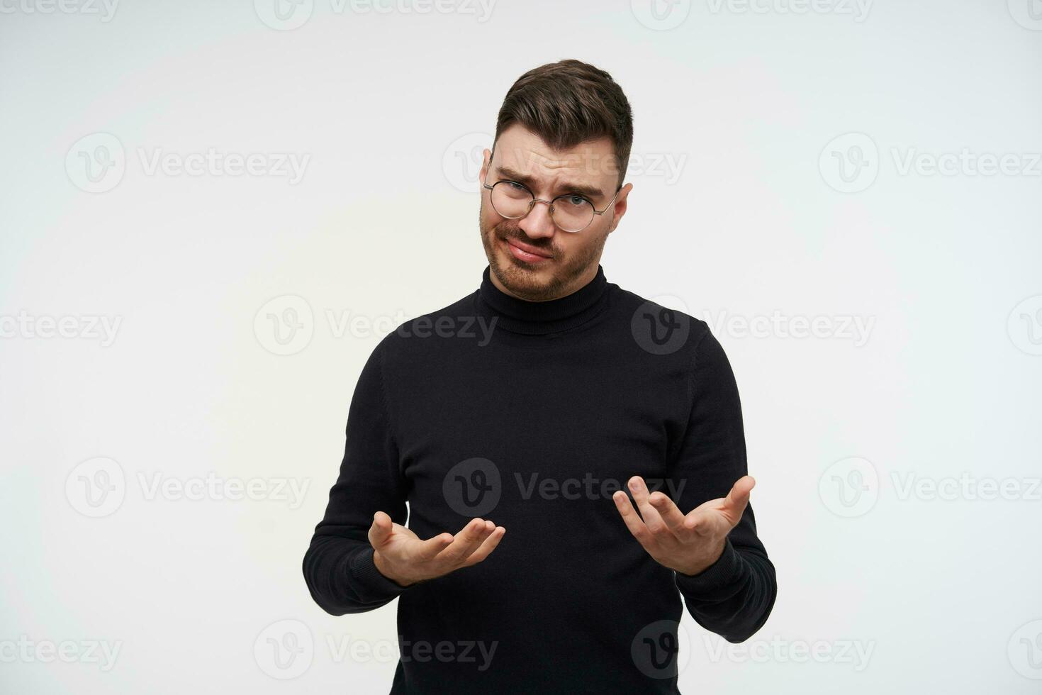 verwirrt jung ziemlich bärtig Brünette Kerl gekleidet im schwarz Rollkragen Verdrehen seine Mund während suchen verwirrt beim Kamera und erziehen Hände, posieren Über Weiß Hintergrund foto