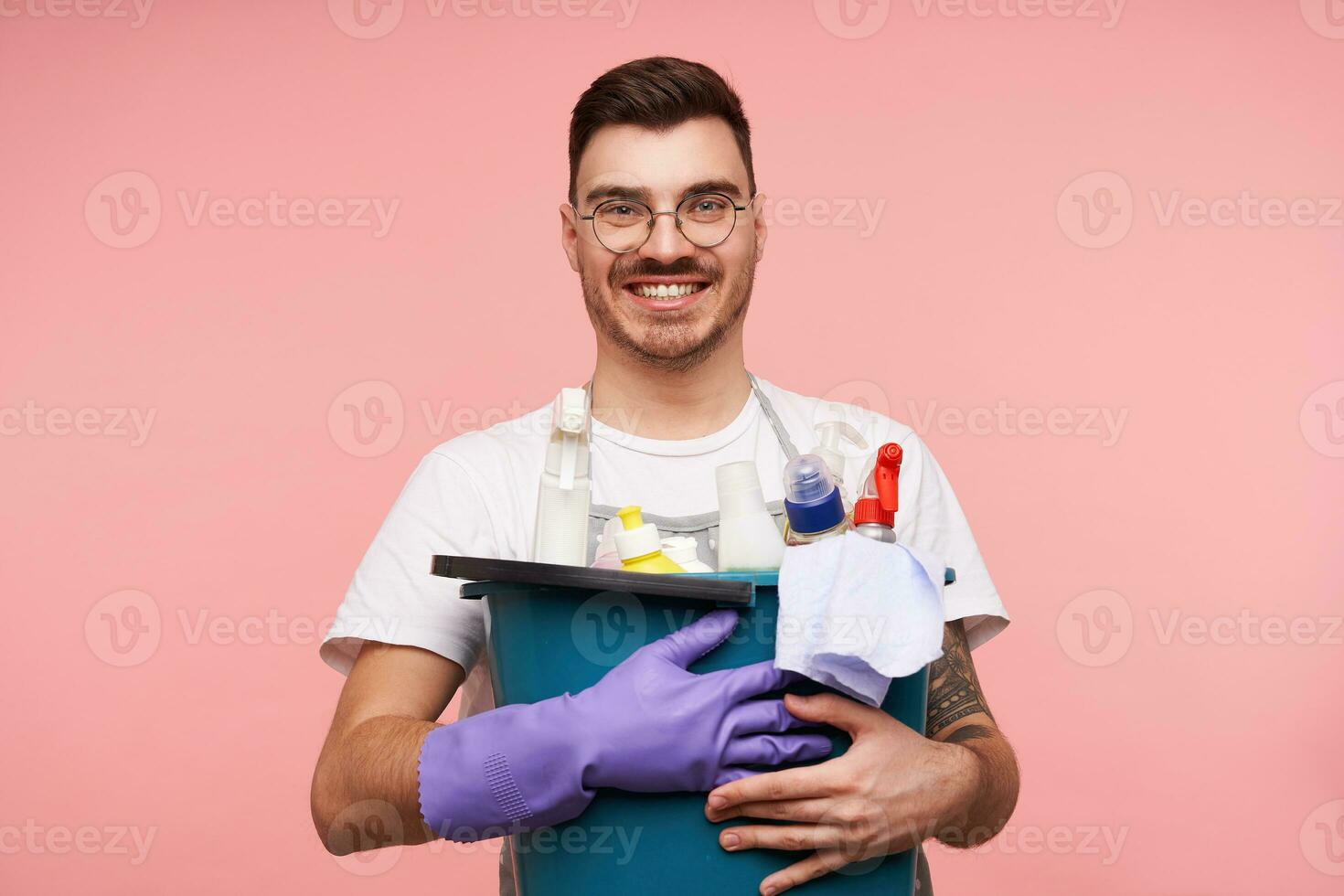 heiter jung attraktiv Brünette männlich mit kurz Haarschnitt lächelnd weit beim Kamera während behalten Busket mit Haushalt Chemikalien im angehoben Hände, isoliert Über Rosa Hintergrund foto