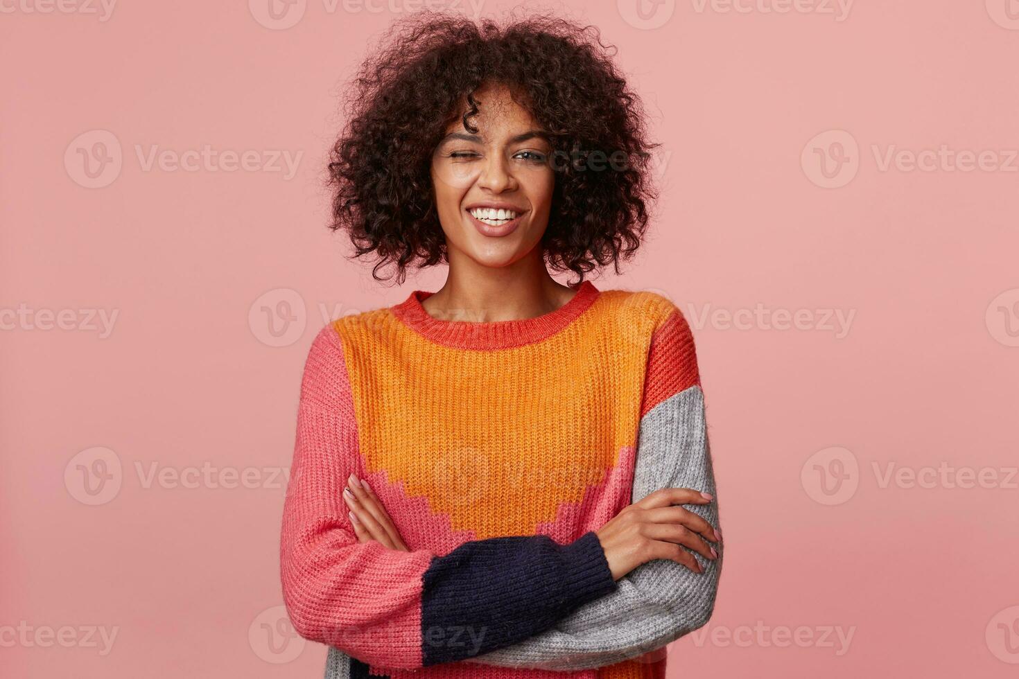 Foto von glamourös charismatisch attraktiv afrikanisch amerikanisch Mädchen mit afro Frisur sieht aus mit Freude, mit glücklich lächeln, Stand mit Waffen gekreuzt, zwinkert, hat Spaß, im bunt lange Ärmel, isoliert auf Rosa