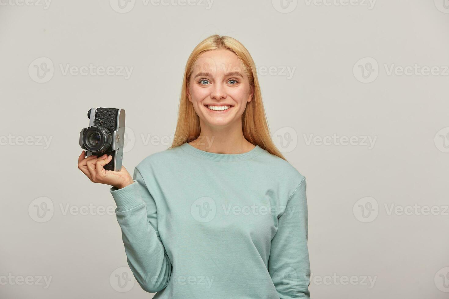 schön Mädchen Fotograf sieht aus glücklich lächelnd, halten ein retro Jahrgang Foto Kamera im einer Hand, bekommt Vergnügen von das Schießen Verfahren, tragen Blau beiläufig Sweatshirt, Über grau Hintergrund