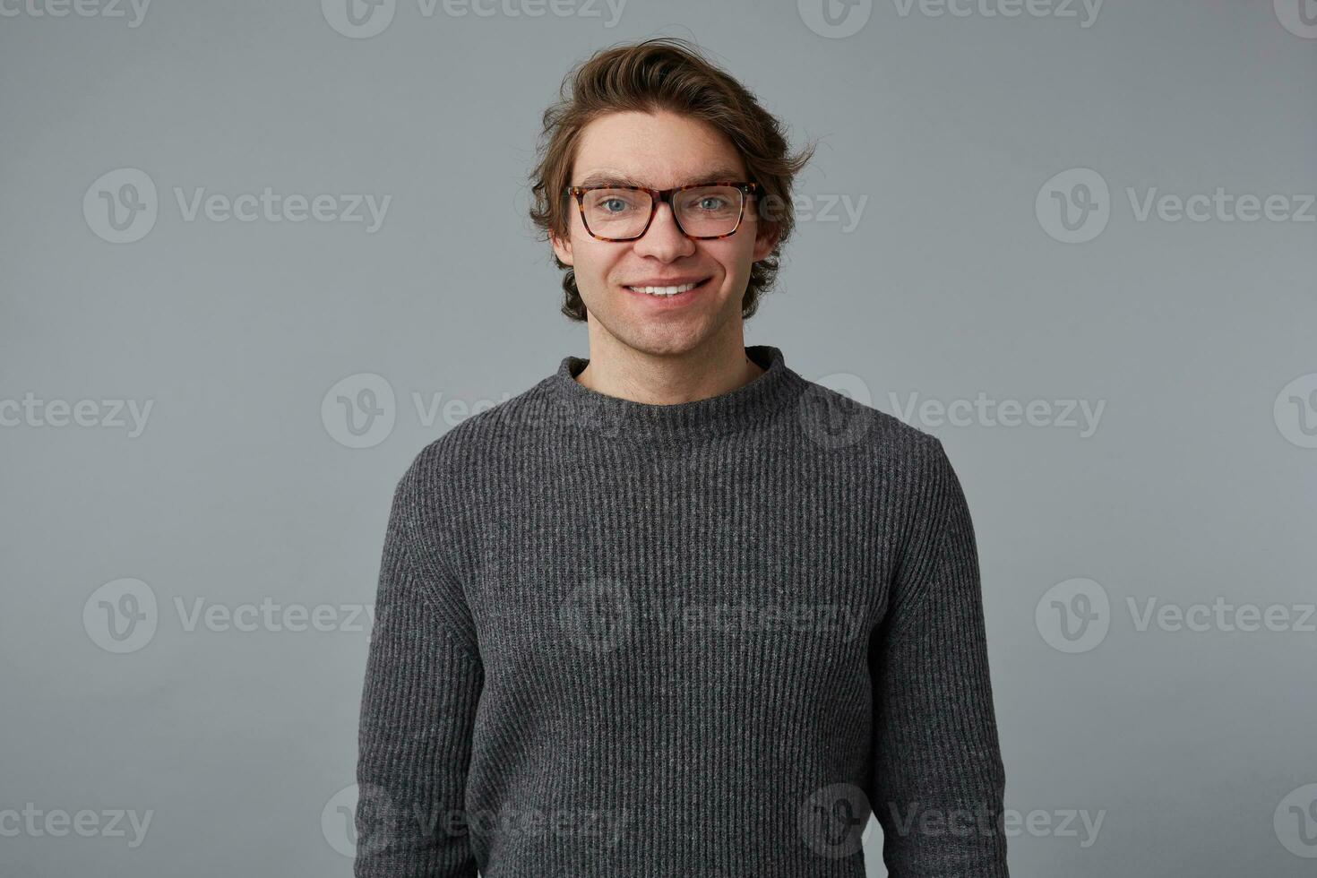 Foto von jung heiter gut aussehend Kerl mit Brille trägt im grau Pullover, steht Über grau Hintergrund und breit lächelt, sieht aus glücklich und froh.