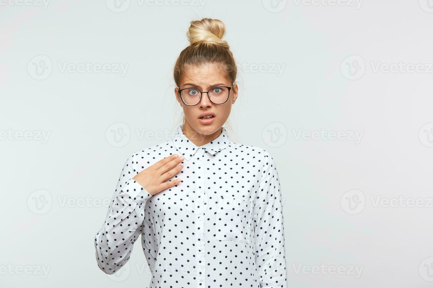 Nahansicht von unzufrieden verärgert jung Frau mit Brötchen trägt Polka Punkt Hemd und Brille fühlt sich unzufrieden und Punkte beim Sie selber isoliert Über Weiß Hintergrund foto