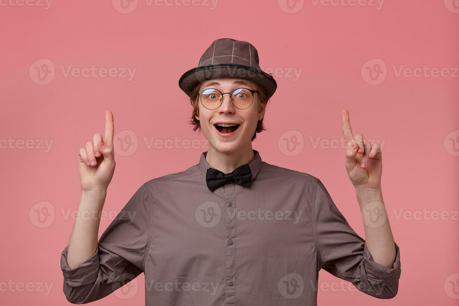 Kerl geöffnet seine Mund im Aufregung, ist überwältigt mit positiv Emotionen Glück Freude zeigen Index Finger nach oben zeichnet Aufmerksamkeit, gekleidet im Hemd Hut Krawatte Brille hat Klammern isoliert auf Rosa foto