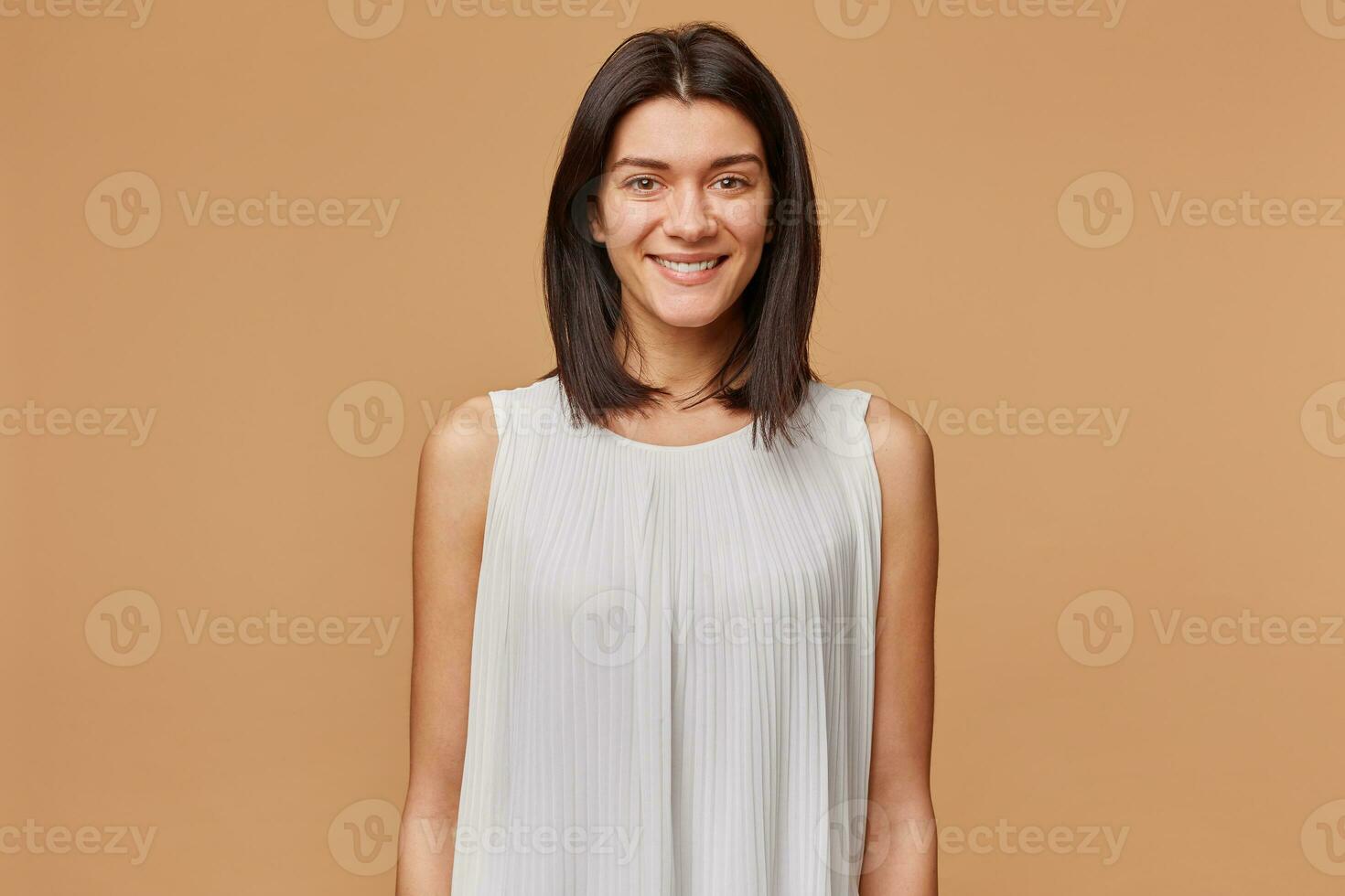 Porträt von ein nett attraktiv schön Mädchen mit dunkel Haar im Weiß Kleid angenehm lächelnd, isoliert Über Beige Hintergrund foto