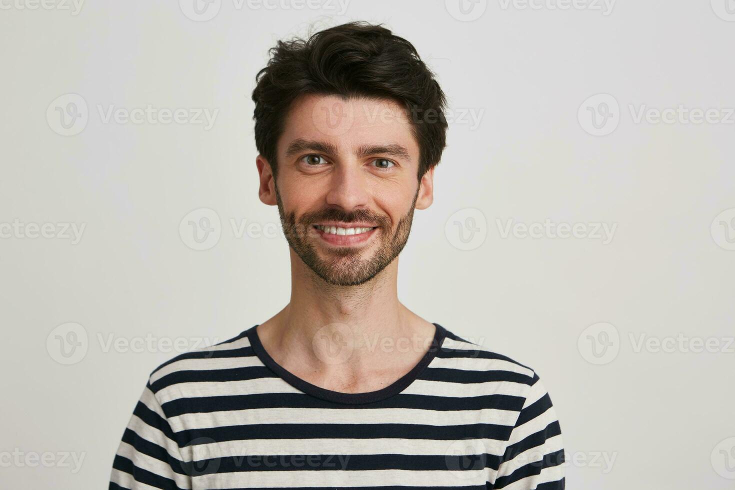 Nahansicht von lächelnd attraktiv bärtig jung Mann trägt gestreift t Hemd fühlt sich glücklich und sieht aus direkt im Kamera isoliert Über Weiß Hintergrund foto