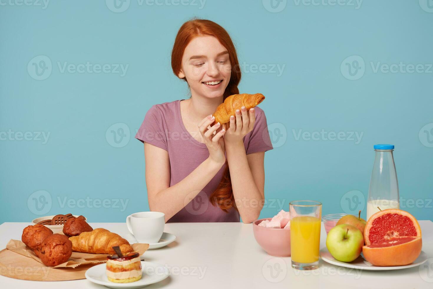 süß rothaarig Mädchen mit ein flechten sitzt beim ein Tisch, hat Frühstück, mit Aufregung Vergnügen sieht aus beim Croissant, Gebäck auf das Tabelle und frisch Obst, auf ein Blau Hintergrund foto