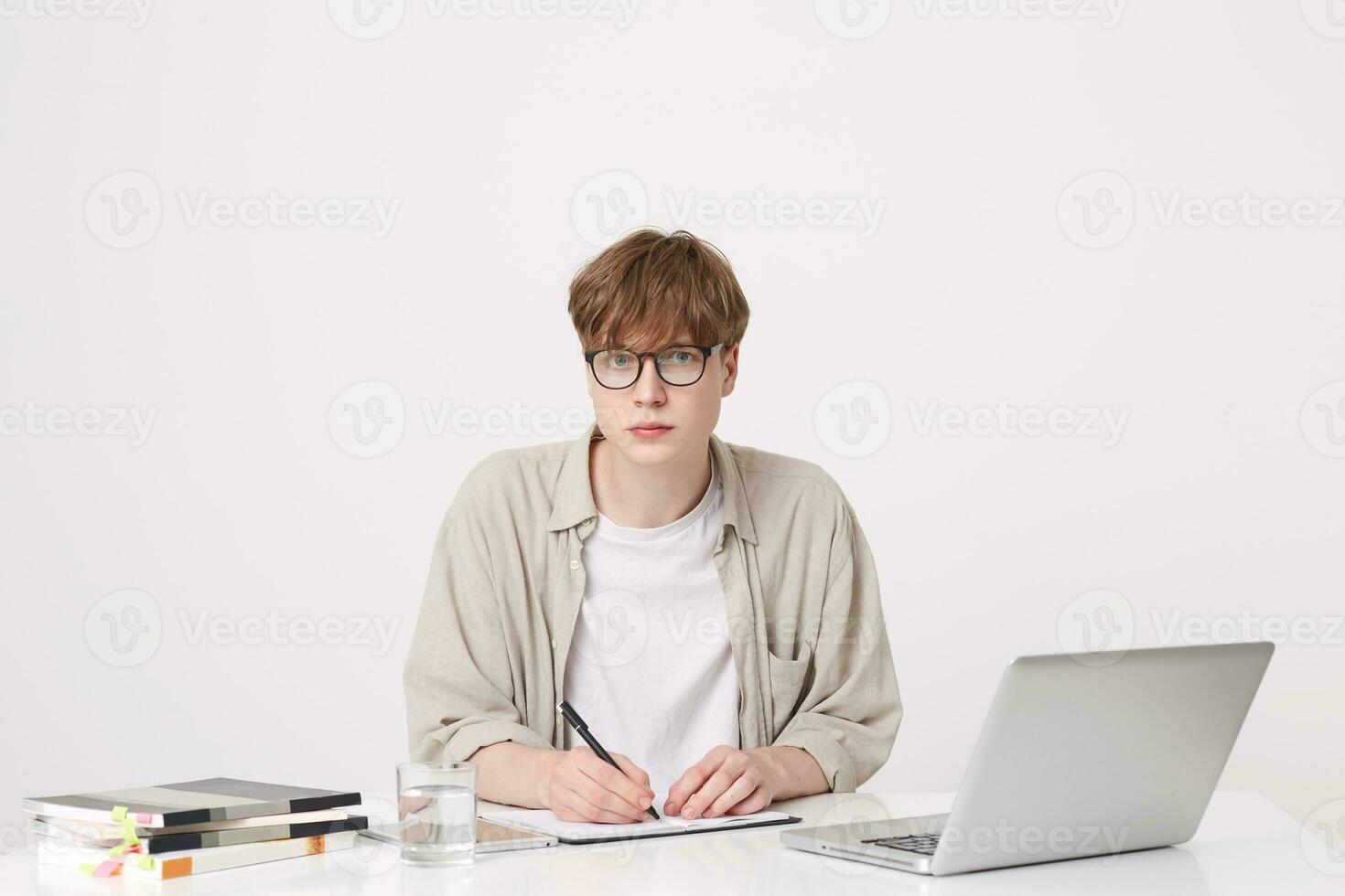 Nahansicht von ernst gut aussehend jung Mann Schüler trägt Beige Hemd und Brille Sitzung mit Laptop Computer und Notizbücher beim das Tabelle und Schreiben isoliert Über Weiß Hintergrund foto