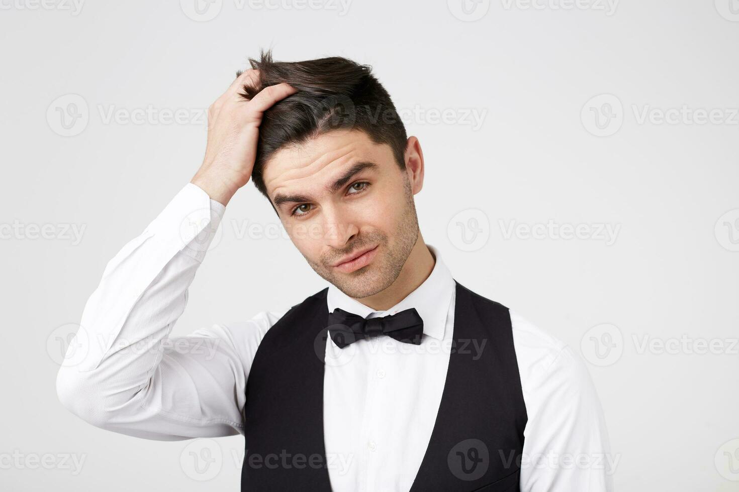 attraktiv Brunert Kerl mit Licht Borsten, smarty gekleidet spielerisch richtet sich auf Haare, seine Kopf ist leicht gekippt, sieht aus mit ein charmant, kokett, verzaubert aussehen in das Kamera, vorbei Weiß Hintergrund foto