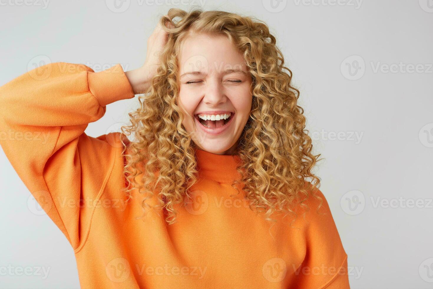 heiter sorglos froh lockiges Haar blond hält ihr Hand in der Nähe von Kopf geschlossen ihr Augen von Vergnügen, lacht glücklich, gekleidet im ein warm Orange Übergröße Pullover, isoliert auf Weiß Hintergrund foto