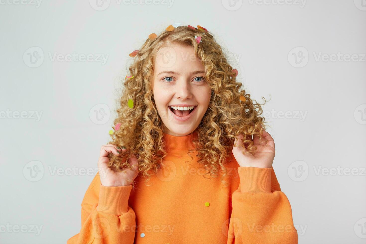 ein Zeit zu haben Spaß. Porträt von ein sehr glücklich Mädchen im Orange Sweatshirt berührt Theaterstücke mit ihr lockig Haar, lächelnd isoliert auf ein Weiß Hintergrund foto