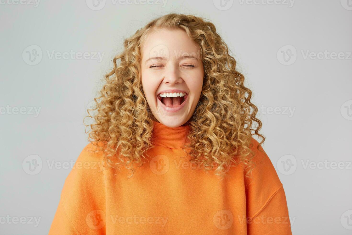 belastbar glücklich heiter lockiges Haar blond geschlossen ihr Augen mit Vergnügen, genießt das Moment Lachen, tragen ein warm Orange Übergröße Pullover, isoliert auf Weiß Hintergrund foto