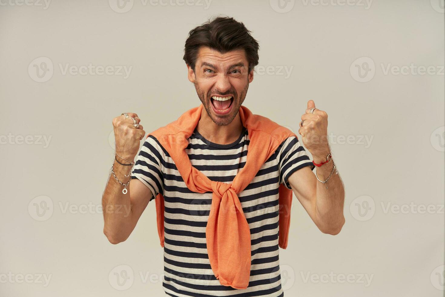 aufgeregt Mann mit Brünette Haar und Borste. tragen gestreift T-Shirt und Orange Sweatshirt gebunden auf Schultern. ballen Fäuste. hat Armbänder und Ringe. Aufpassen beim das Kamera isoliert Über grau Hintergrund foto