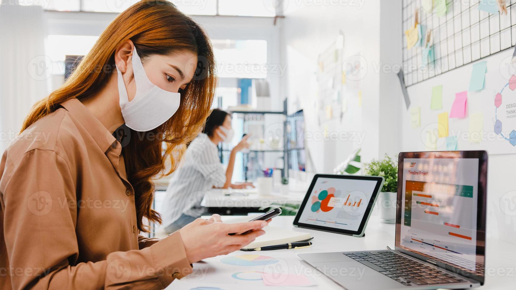 Unternehmerin aus Asien, die Gesichtsmaske für soziale Distanzierung in einer neuen normalen Situation zur Virusprävention trägt, während sie Laptop und Telefon bei der Arbeit im Büro verwendet. Lebensstil nach dem Corona-Virus. foto