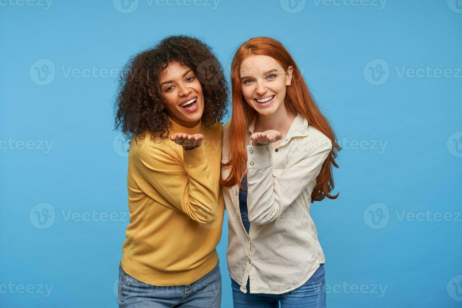 glücklich attraktiv jung Freundinnen umarmen jeder andere und lächelnd breit während suchen fröhlich zu Kamera, behalten ihr Palmen angehoben während posieren Über Blau Hintergrund foto