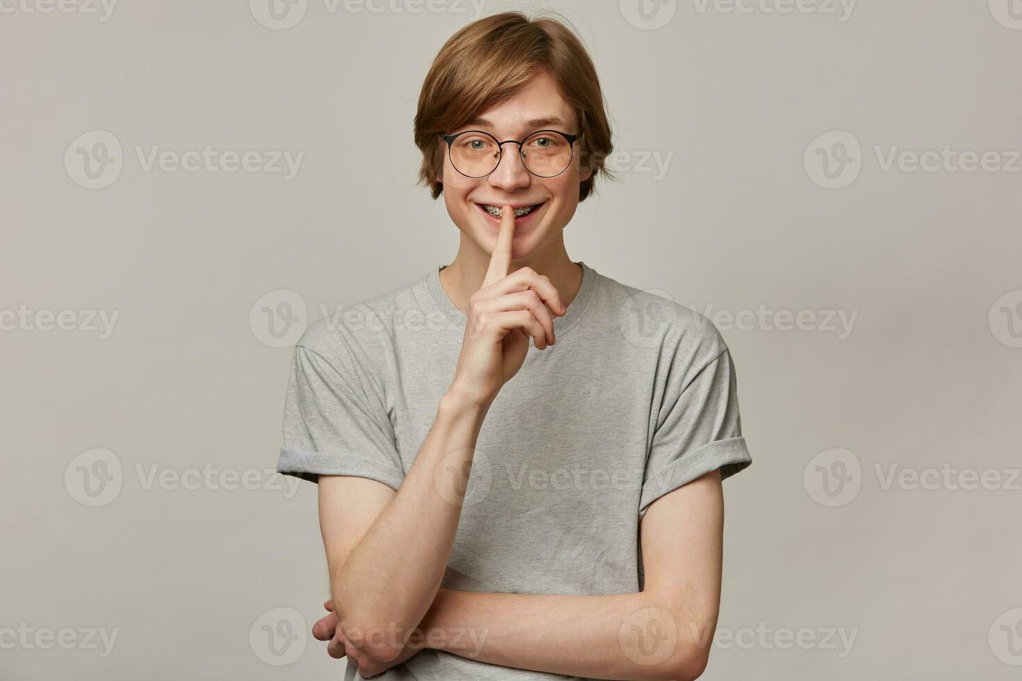 Teenager Kerl, glücklich suchen Mann mit blond Haar. tragen grau T-Shirt, Brille und hat Zahnspange. Menschen und Emotion Konzept. zeigen Stille unterzeichnen. Aufpassen beim das Kamera isoliert Über grau Hintergrund foto