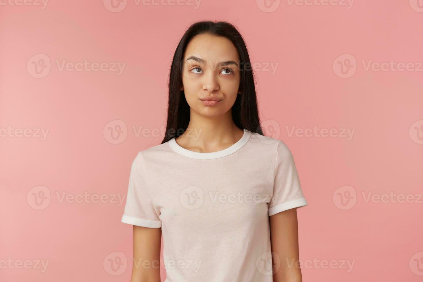 Teenager Mädchen, genervt suchen Frau mit dunkel lange Haar. tragen Weiß T-Shirt. Menschen und Emotion Konzept. Rollen ihr Augen und geschürzt Lippen. Aufpassen oben beim Kopieren Raum, isoliert Über Rosa Hintergrund foto