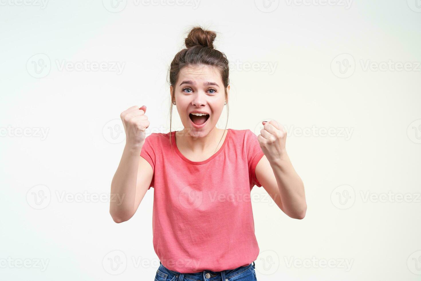 froh jung ziemlich Brünette weiblich mit Brötchen Frisur erziehen glücklich Fäuste während suchen aufgeregt beim Kamera mit geöffnet Mund, Stehen Über Weiß Hintergrund foto