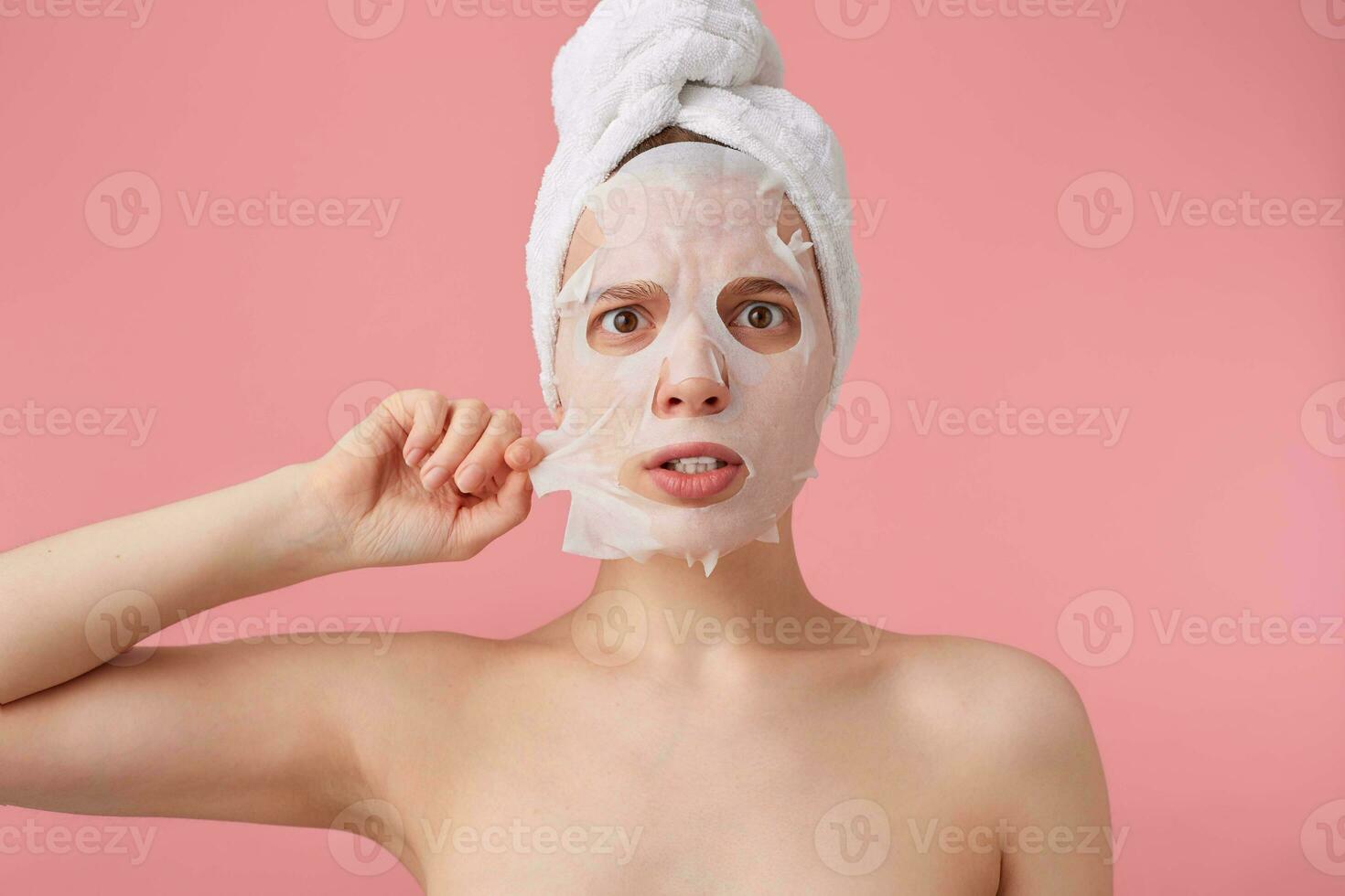 schließen oben von jung schockiert Frau mit ein Handtuch auf ihr Kopf nach Dusche, versuchen zu entfernen das Stoff Maske von das Gesicht, steht Über Rosa Hintergrund. foto