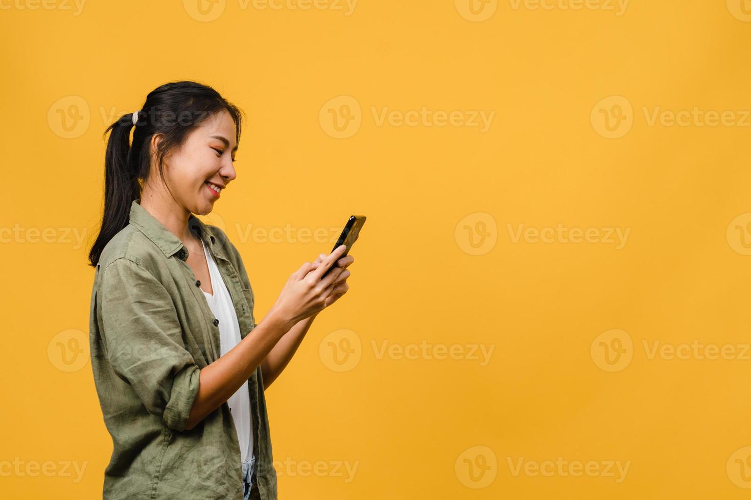 junge asiatische dame, die telefon mit positivem ausdruck verwendet, breit lächelt, in lässiger kleidung gekleidet fühlt sich glücklich und steht einzeln auf gelbem hintergrund. glückliche entzückende frohe frau freut sich über erfolg. foto