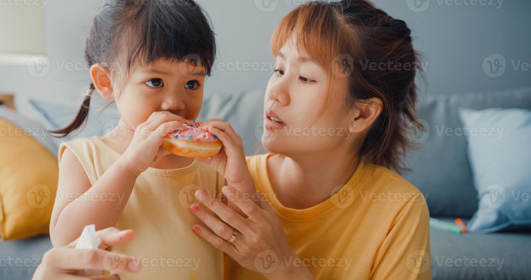 Fröhliche, fröhliche Asien-Familienmutter und Kleinkindmädchen, die Donuts essen und Spaß haben, entspannen Sie sich auf der Couch im Wohnzimmer im Haus. Zeit miteinander verbringen, soziale Distanz, Quarantäne für Coronavirus. foto