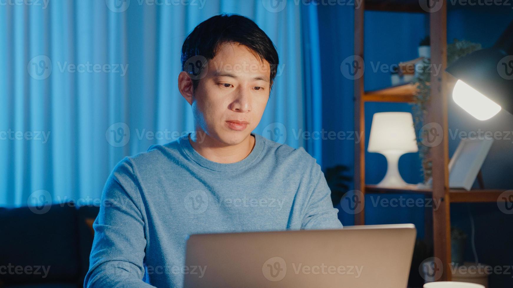 asiatischer freiberuflicher geschäftsmann konzentriert sich auf das arbeiten auf laptop-computer online fern von der firma auf dem schreibtisch im wohnzimmer zu hause überstunden nachts, arbeiten von zu hause während des covid-19-pandemiekonzepts. foto