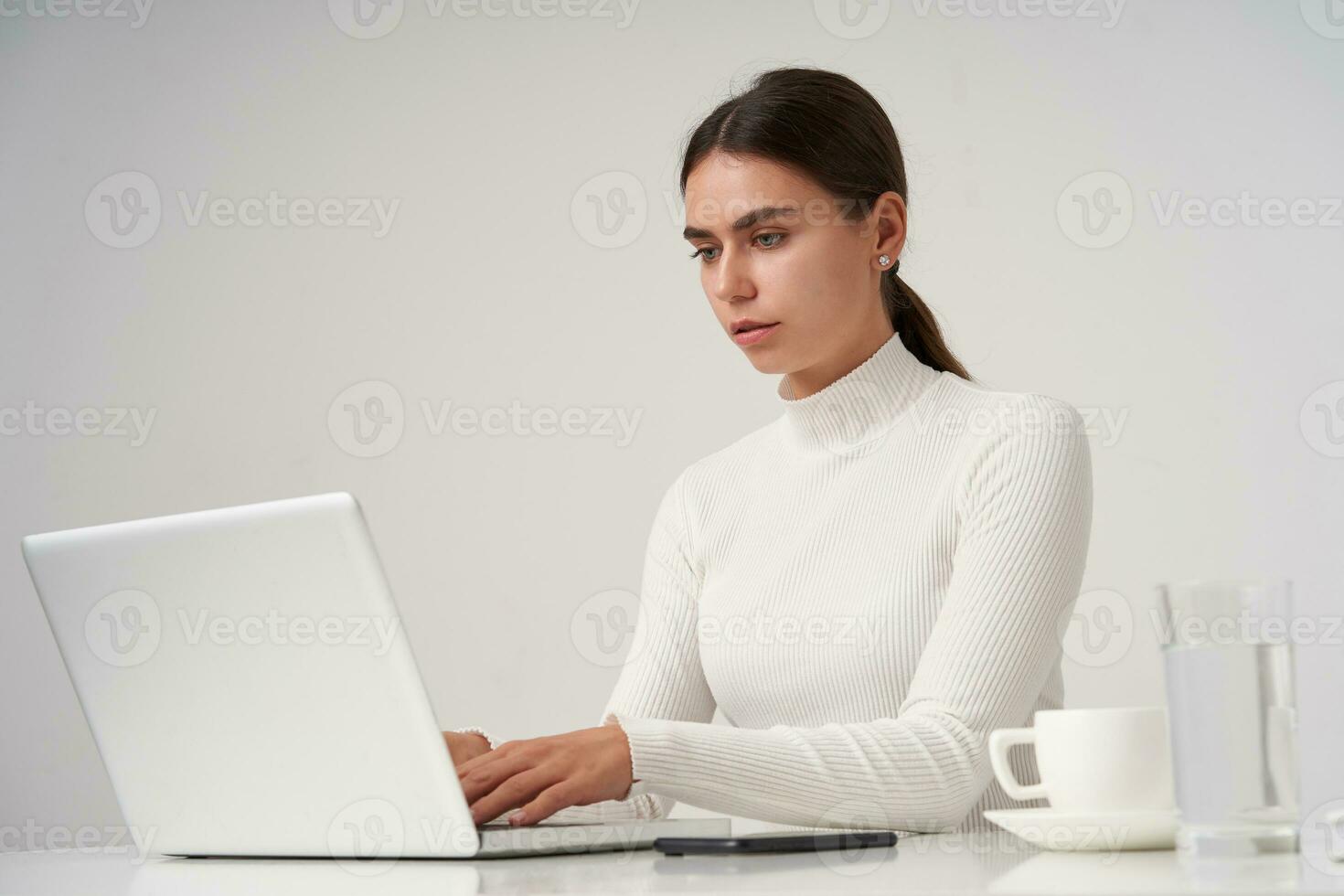 ernst jung schön Brünette weiblich suchen beim Bildschirm mit konzentriert Gesicht während Tippen Text auf Klaviatur, Sitzung beim Tabelle Über Weiß Hintergrund foto