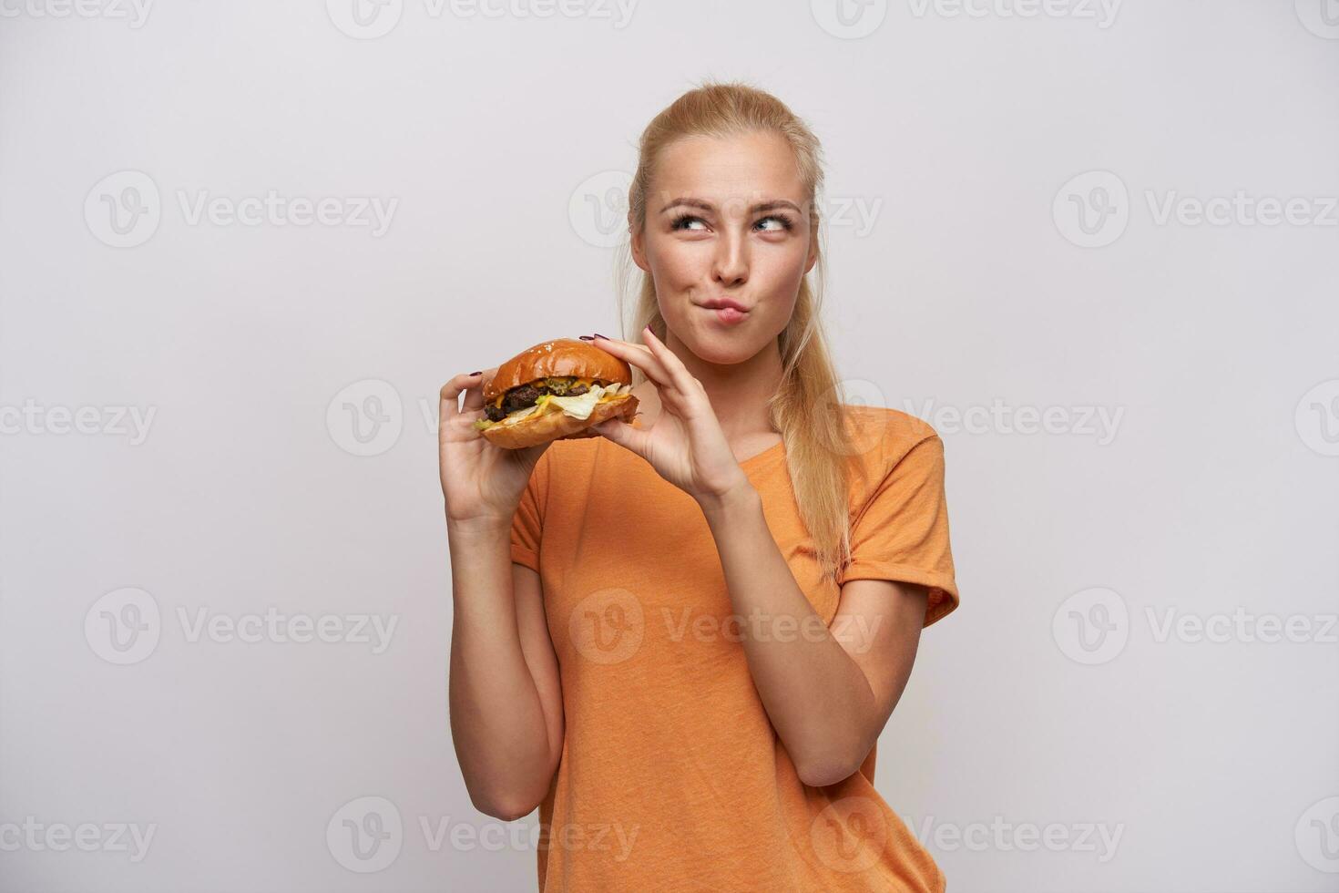 Studio Schuss von positiv jung ziemlich lange behaart blond weiblich Essen frisch Burger und suchen zweifelhaft beiseite, gekleidet im Orange T-Shirt während Stehen Über Weiß Hintergrund foto