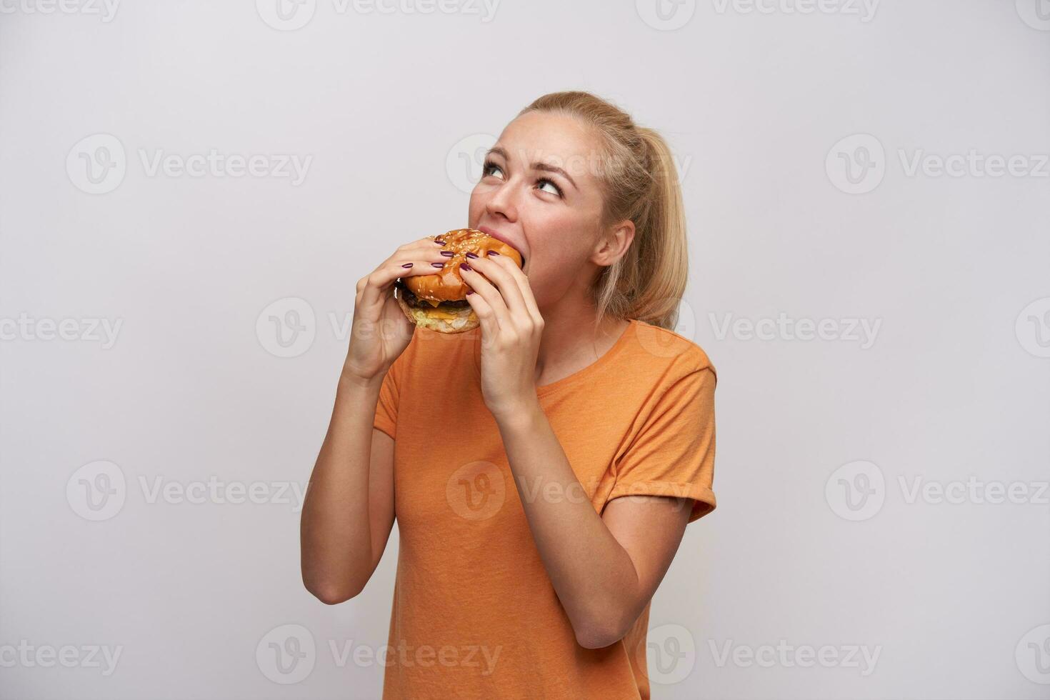 Innen- Foto von zufrieden jung lange behaart blond Frau mit Pferdeschwanz Frisur tragen beiläufig Kleider während posieren Über Weiß Hintergrund, Essen lecker Hamburger und suchen positiv beiseite