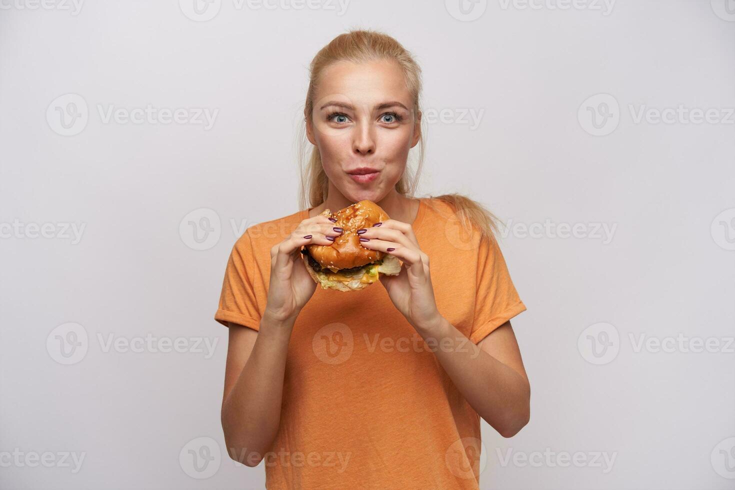 Innen- Foto von attraktiv jung hungrig blond Dame mit Pferdeschwanz Frisur Kauen köstlich Hamburger und suchen aufgeregt beim Kamera, gekleidet im Orange T-Shirt Über Weiß Hintergrund