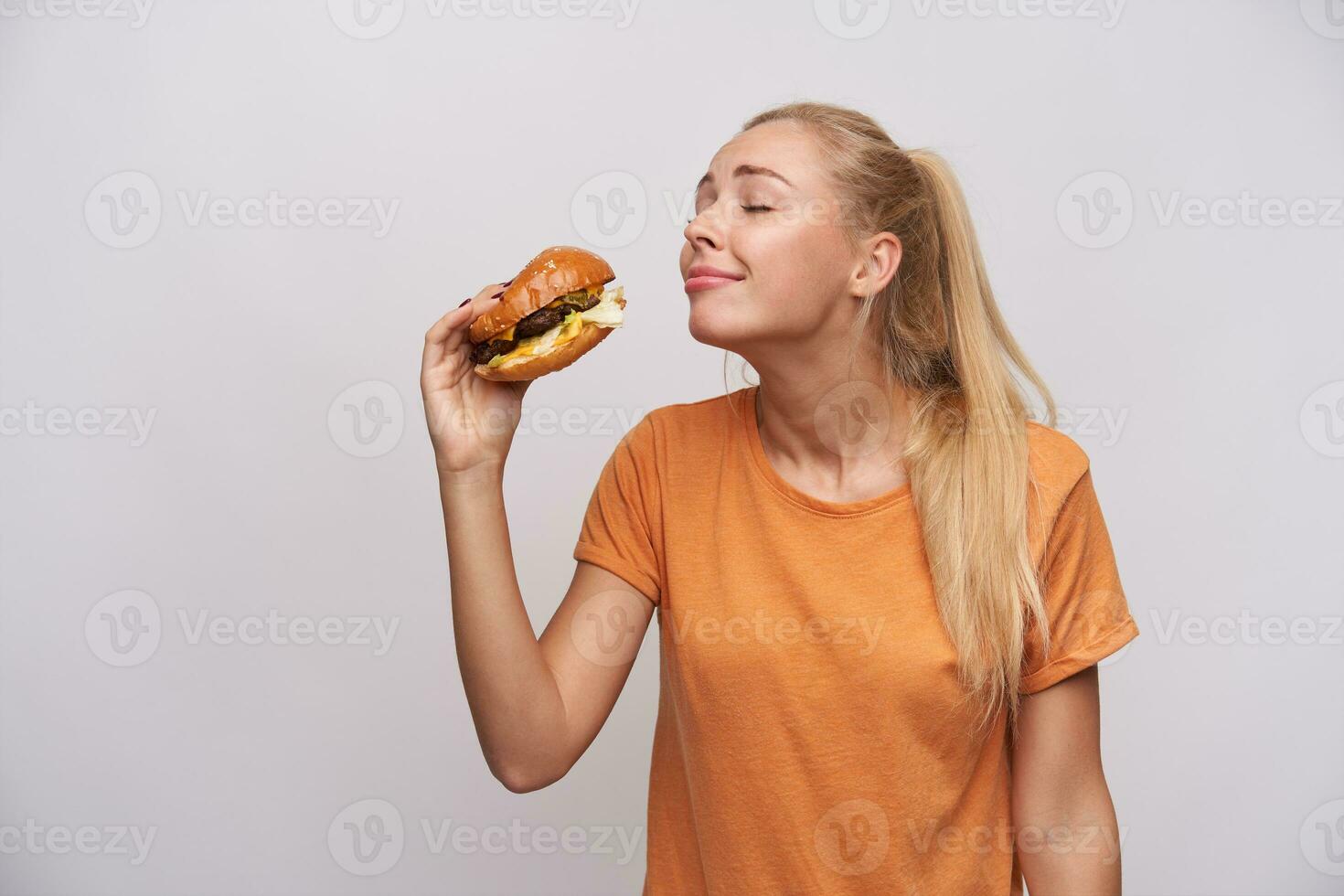 Studio Schuss von positiv jung blond Frau mit Pferdeschwanz Frisur riechen Hamburger im ihr Hand mit großartig Vergnügen und Vorgeschmack köstlich Mahlzeit, Stehen gegen Weiß Hintergrund foto