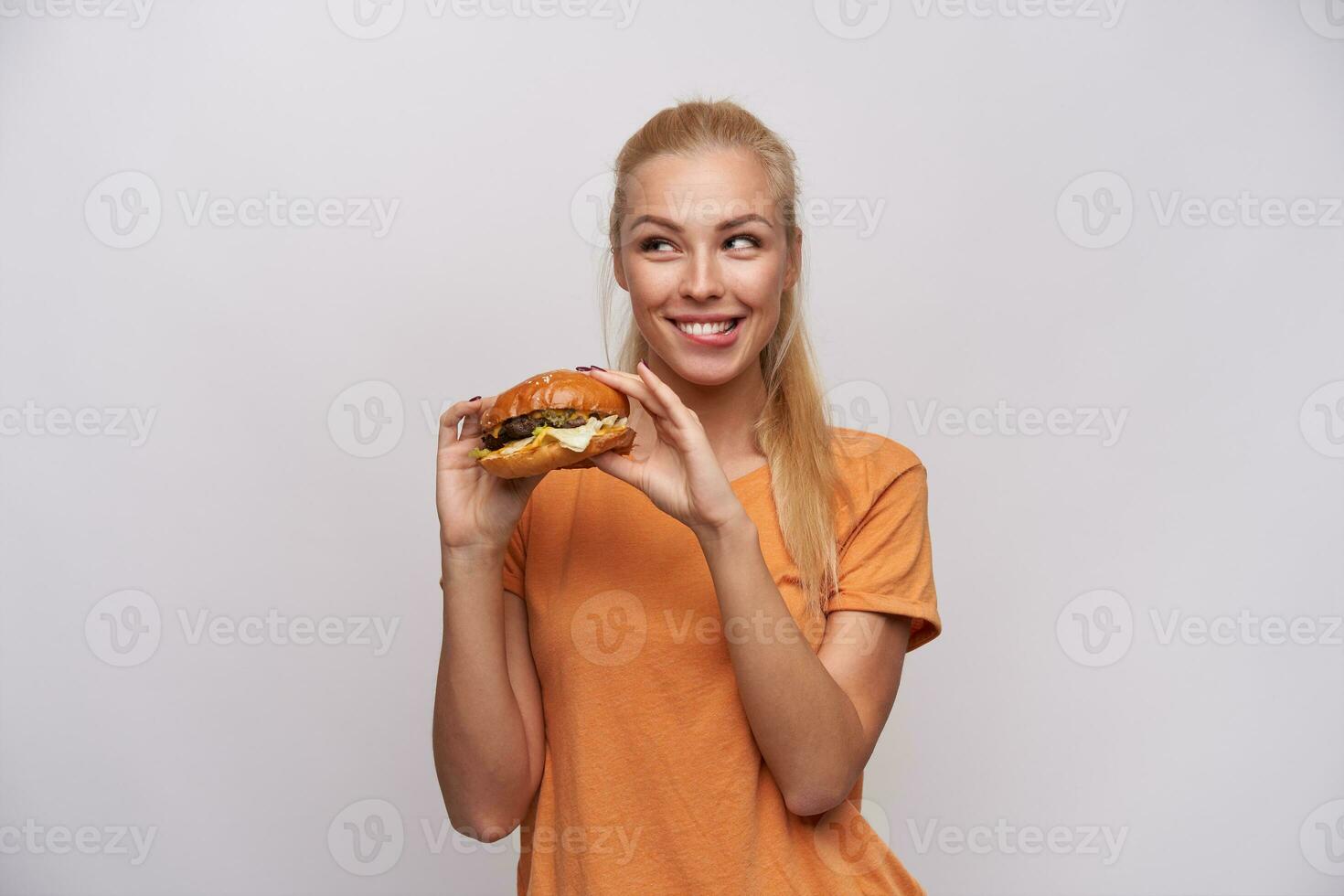 heiter jung schön blond Frau mit beiläufig Frisur halten Cheeseburger im angehoben Hände und suchen listig beiseite mit breit lächeln, isoliert Über Weiß Hintergrund foto