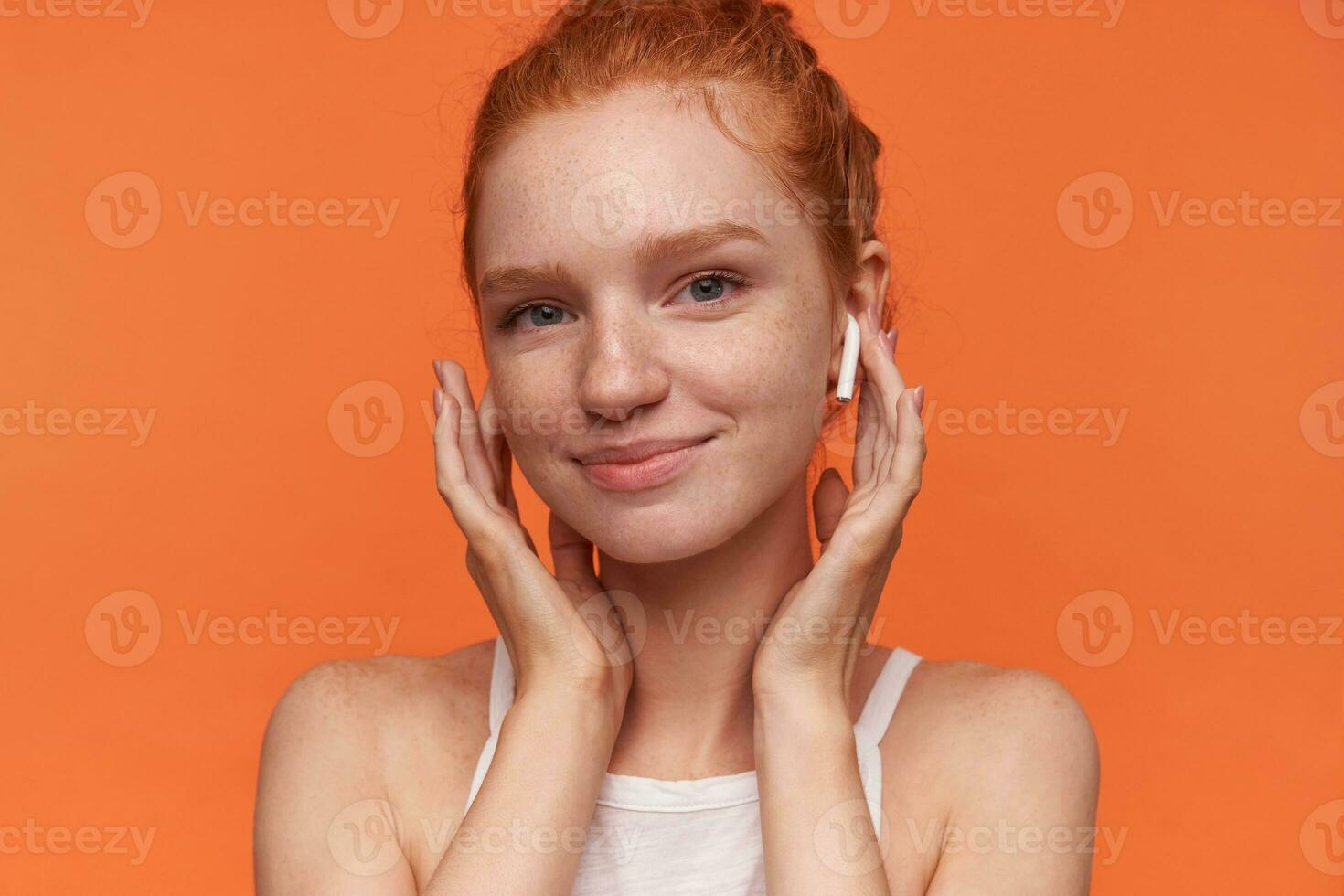 Porträt von jung ziemlich Frau tragen foxy Brötchen Frisur, halten Hände auf Kopfhörer und lächelnd fröhlich zu Kamera, tragen beiläufig Kleidung, isoliert Über Orange Hintergrund foto