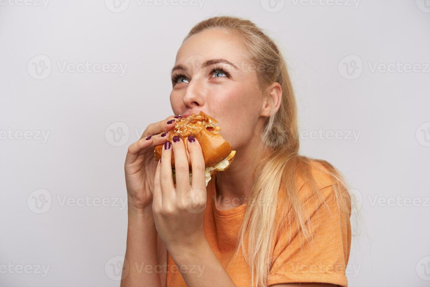 Nahansicht von jung ziemlich blauäugig blond Dame mit beiläufig Frisur behalten groß lecker Burger im ihr Hände und beißen Stück gierig, Stehen gegen Weiß Hintergrund foto