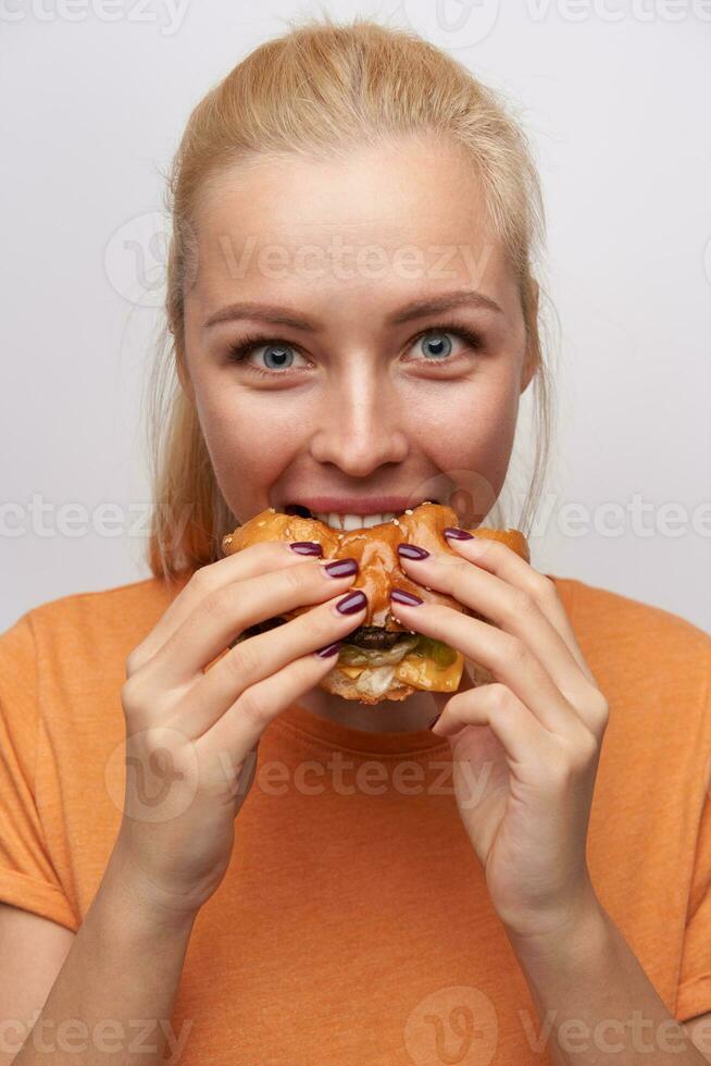 Porträt von aufgeregt jung blauäugig ziemlich blond Frau Essen eifrig ihr Burger und Rundung Augen während suchen freudig beim Kamera, posieren Über Weiß Hintergrund im beiläufig Kleider foto