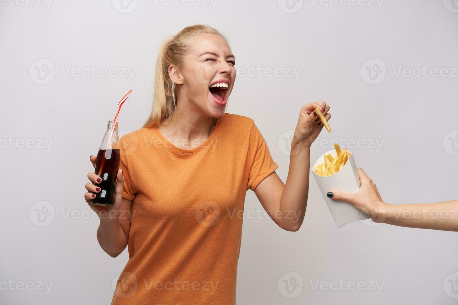 überglücklich attraktiv jung lange behaart blond Frau im beiläufig Kleider Lachen glücklich während nehmen Französisch Fritten von Papier Verpackung und halten Flasche von Limonade, isoliert Über Weiß Hintergrund foto