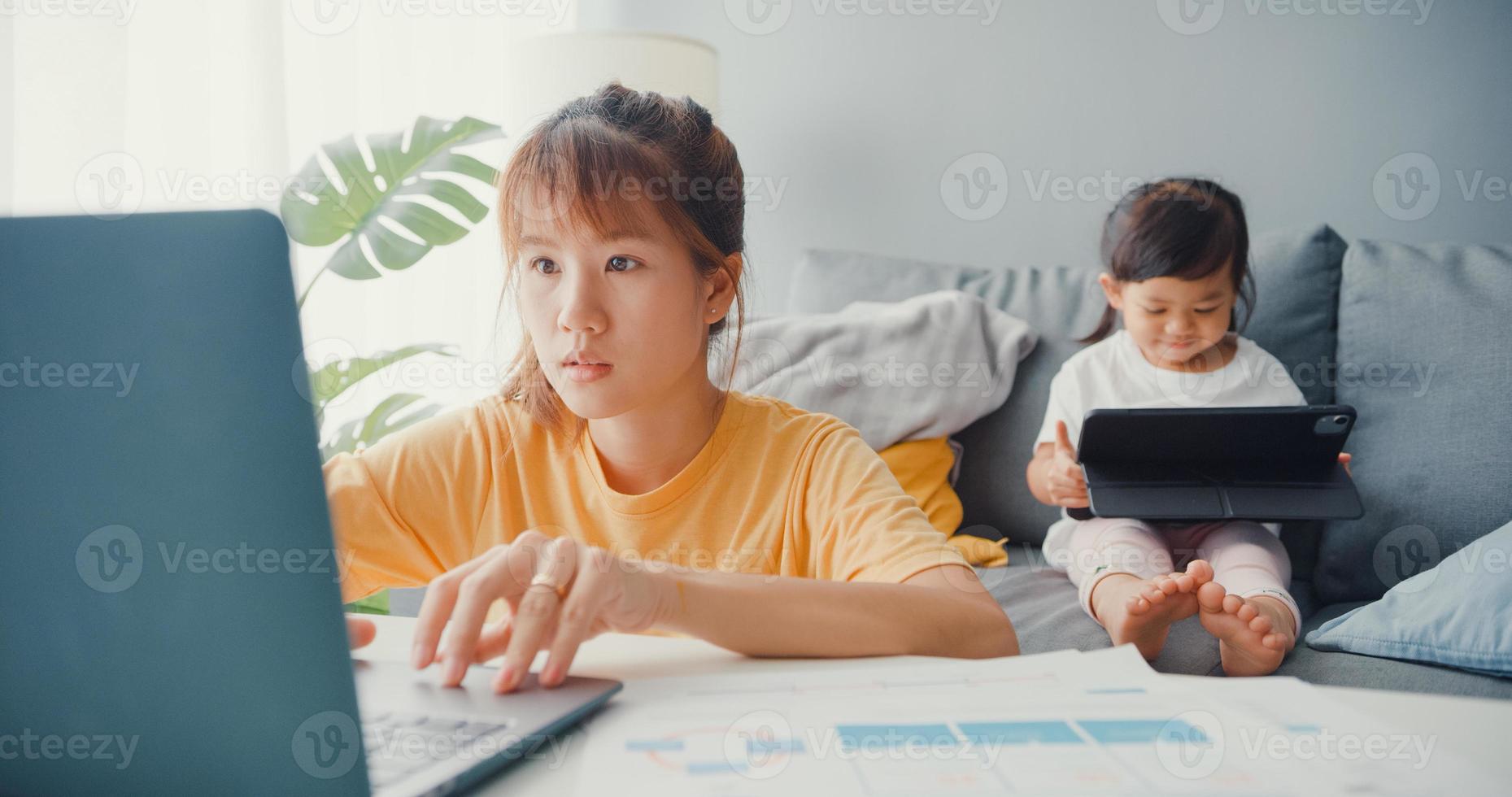 Fröhliche, fröhliche asiatische Familienmutter, die Laptop mit ihrem Kind verwendet, hält Tablet-Interesse-Cartoon auf der Couch, während sie vom Haus im Wohnzimmer arbeitet. soziale Distanzierung, Quarantäne zur Coronavirus-Prävention. foto