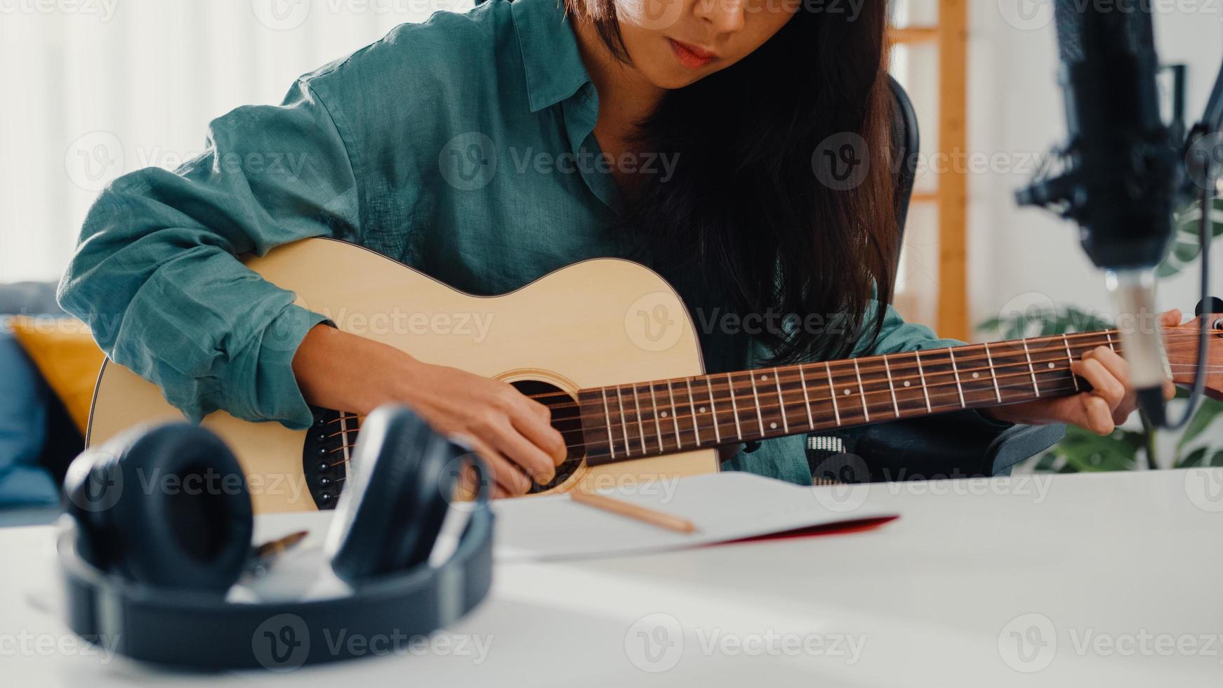 Happy Asia Woman Songwriter spielen Akustikgitarre, hören Song vom Smartphone, denken und schreiben Sie Notizen Songtexte Song in Papier sitzen im Wohnzimmer im Heimstudio. Musikproduktion zu Hause Konzept. foto