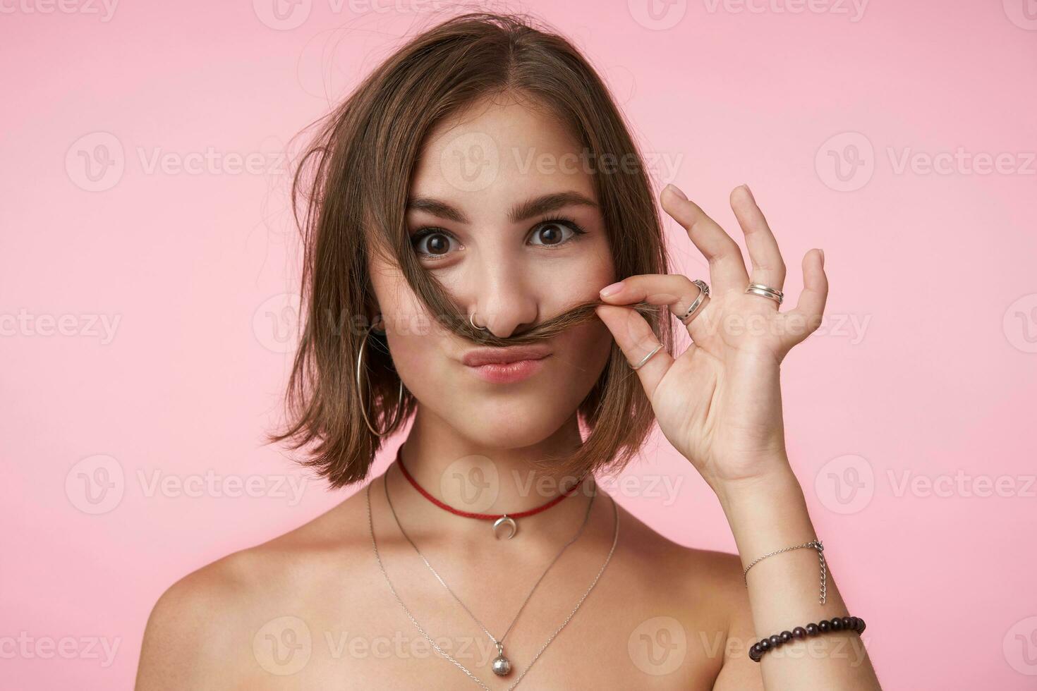 positiv jung kurz behaart Brünette Frau mit natürlich bilden imitieren Schnurrbart mit ihr Haar während Herstellung Spaß, Sein im nett Stimmung während Stehen Über Rosa Hintergrund foto